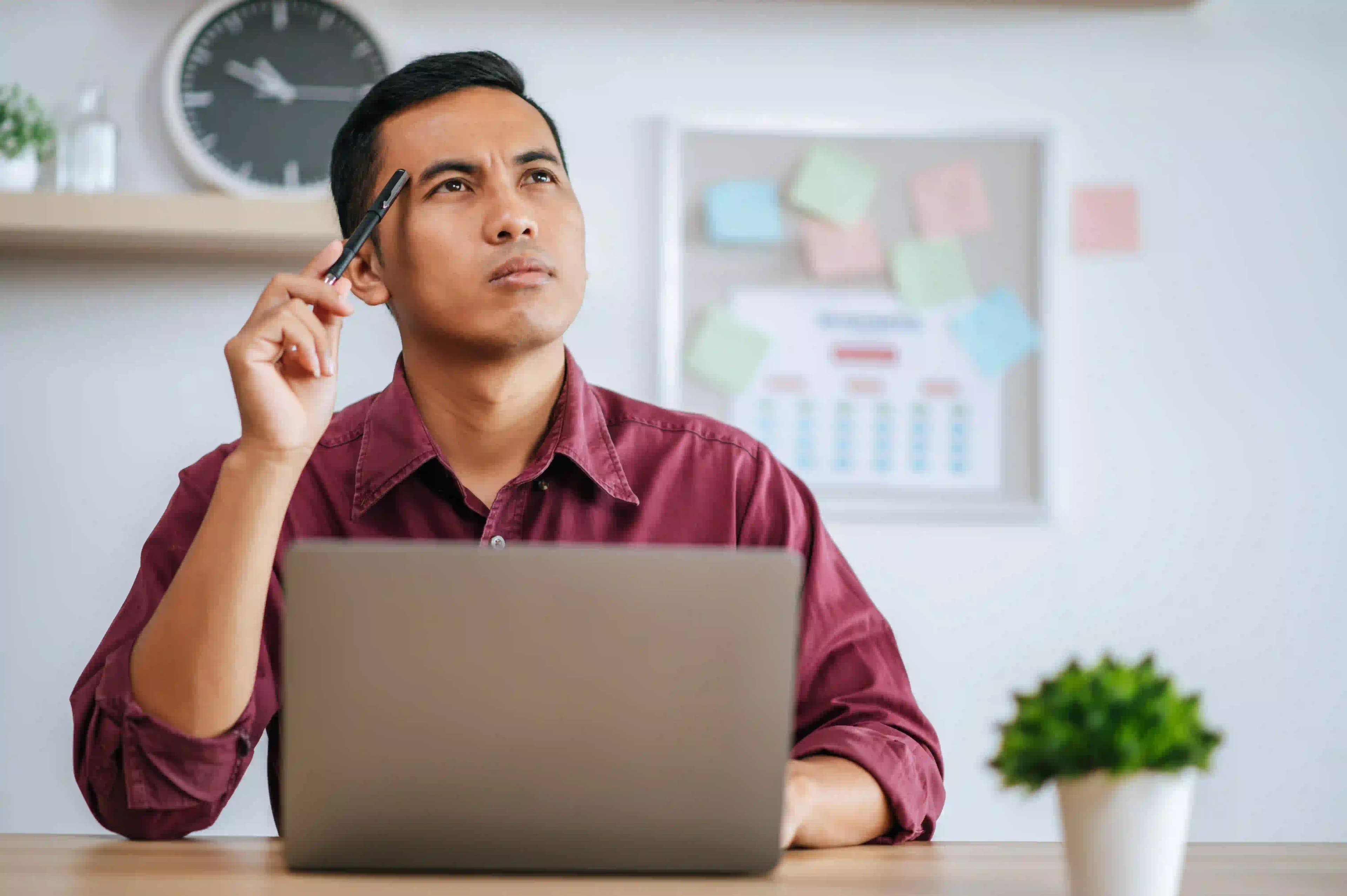 Man at a desk with a laptop, holding a pen and looking thoughtful.